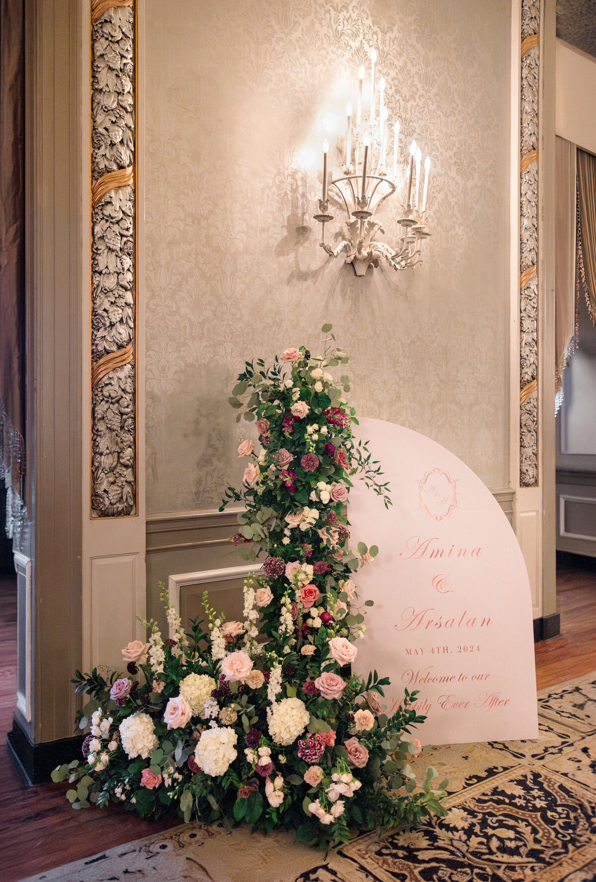 A decorative wedding sign with floral arrangements, featuring roses and greenery, placed on a wooden floor. The wall behind is ornately decorated, and a chandelier hangs above, casting a warm glow over the scene.