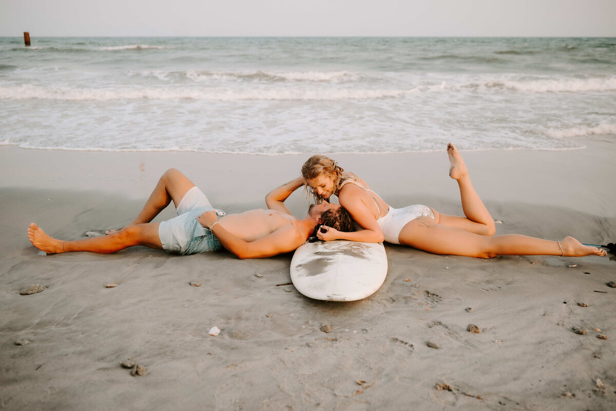 A couple relaxes on sandy beach with surfboard, ocean waves in background. Woman leans on man's chest, conveying a sense of warmth and tranquility.