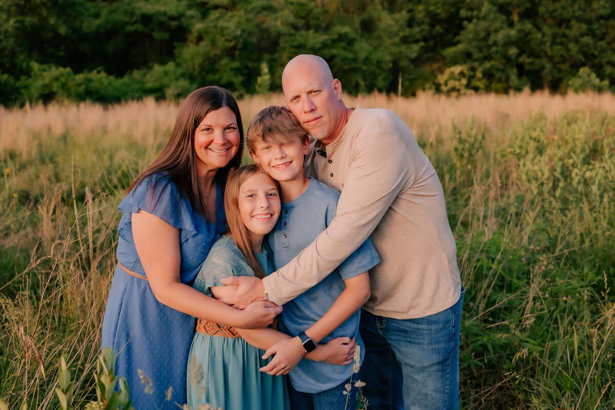 A mom and dad wrap their arms around their two kids pulling them in close to each other for their family photos at sunset.