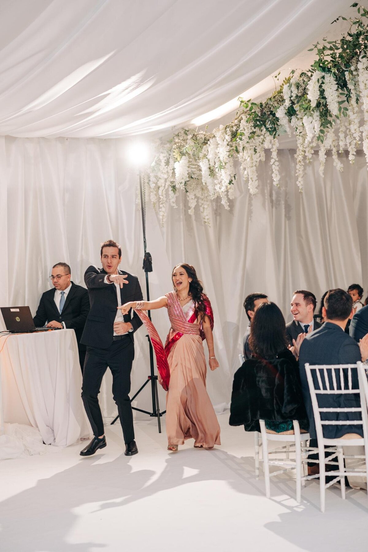 A woman in a pink dress and a man in a suit enthusiastically dancing at a wedding reception while a dj performs.