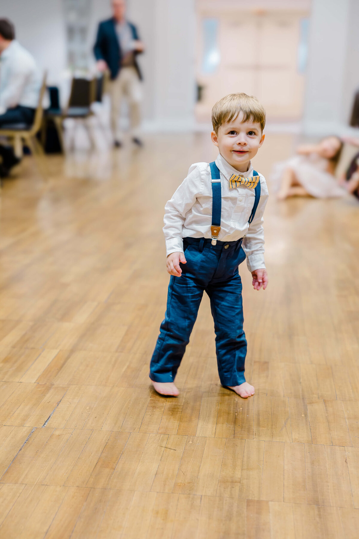 A young boy stands on a wooden floor, dressed in a white shirt, blue suspenders, and blue pants. He is barefoot and indoors. In the background, captured expertly by an event photographer from Fayetteville NC, a person sits on the floor while another person walks gracefully.