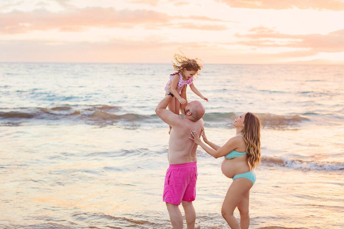 Pregnant mother holds her husband's waist as he lifts their daughter in the air during a fun beach family photoshoot in Wailea