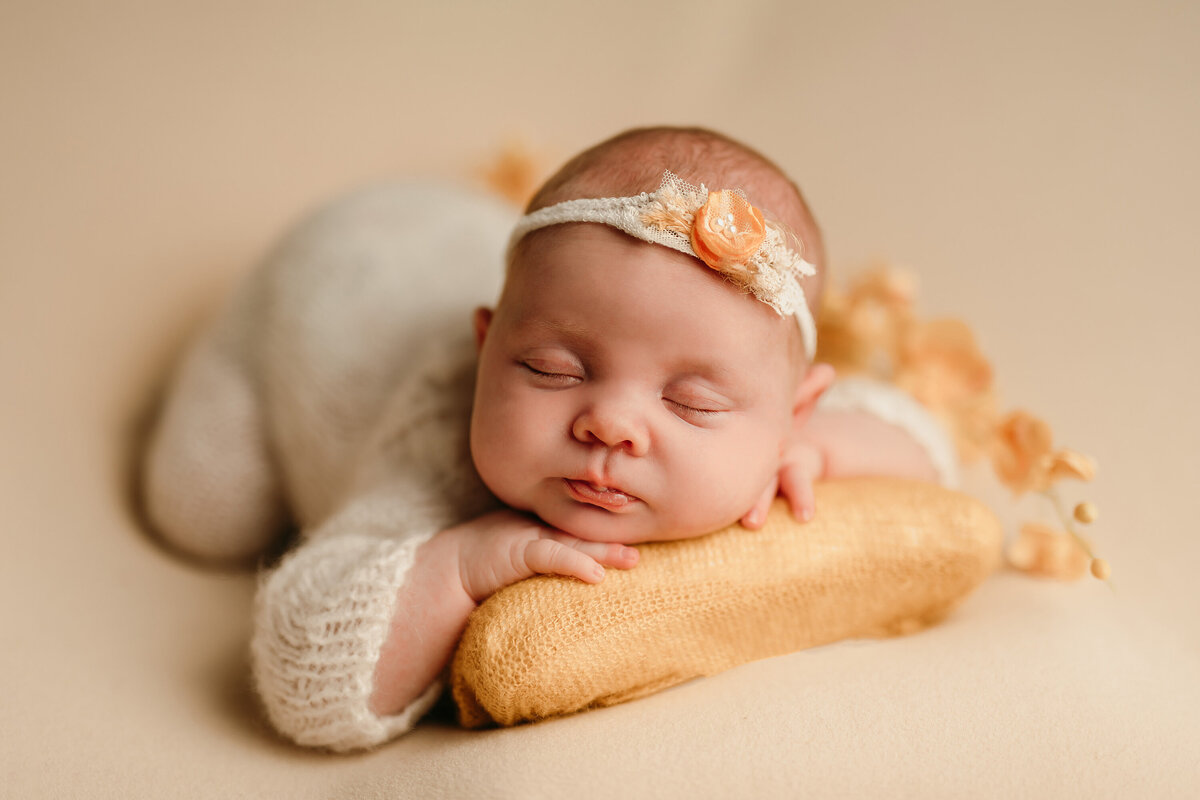 Portrait of newborn baby girl sleeping on her belly with an orange pillow and coordinating floral headband.