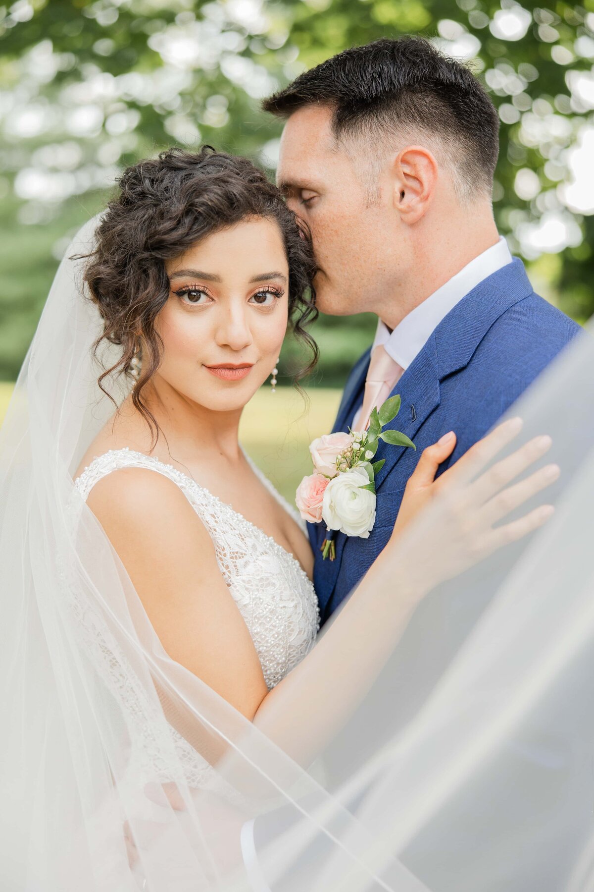 A bride with curly hair and a groom in a blue suit share a tender moment at their wedding in Des Moines, with the bride looking at the camera through her veil.