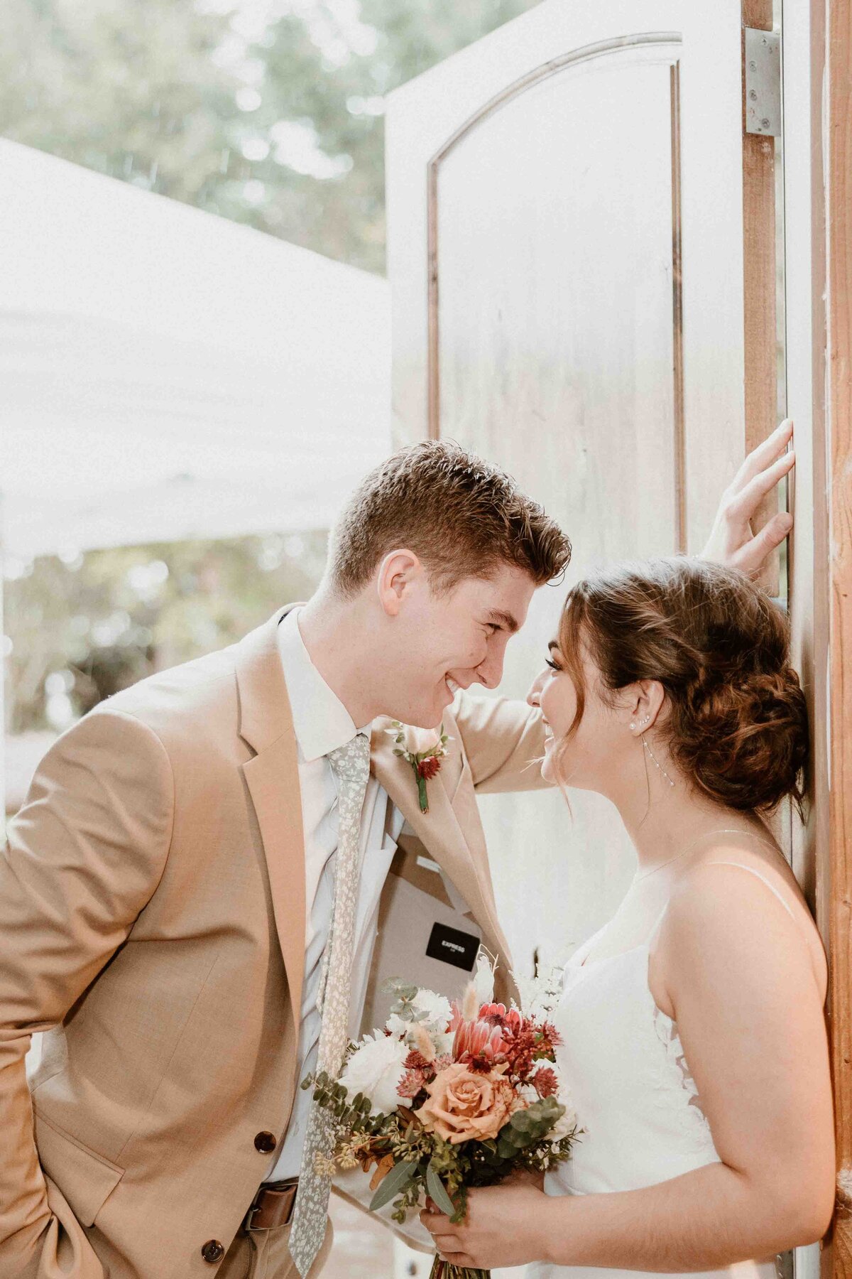 A bride holds her bouquet while leaning on a doorframe with her groom leaning over her