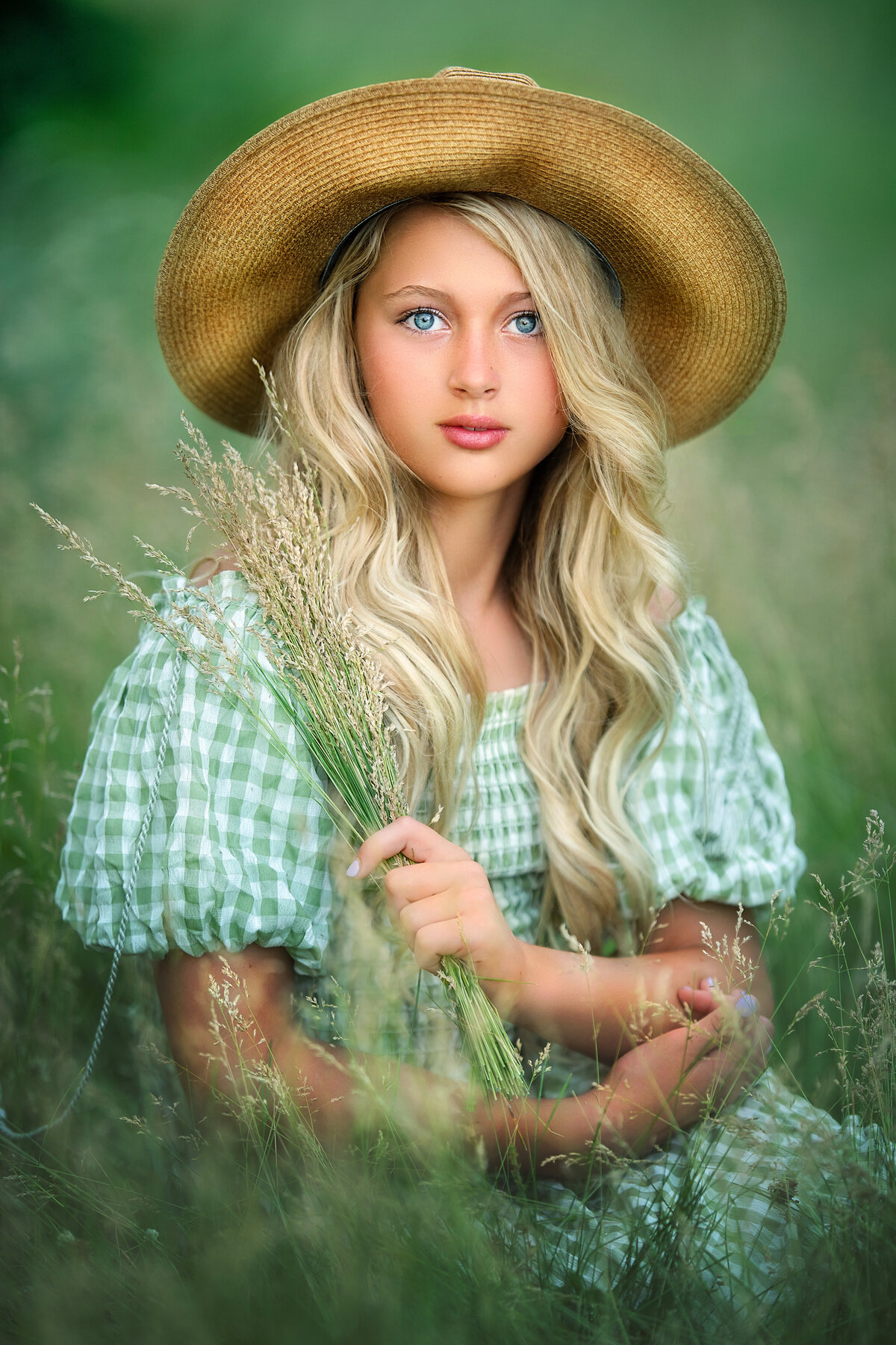 Blond girl with blue eyes and straw hat and green dress sitting in the tall grass.