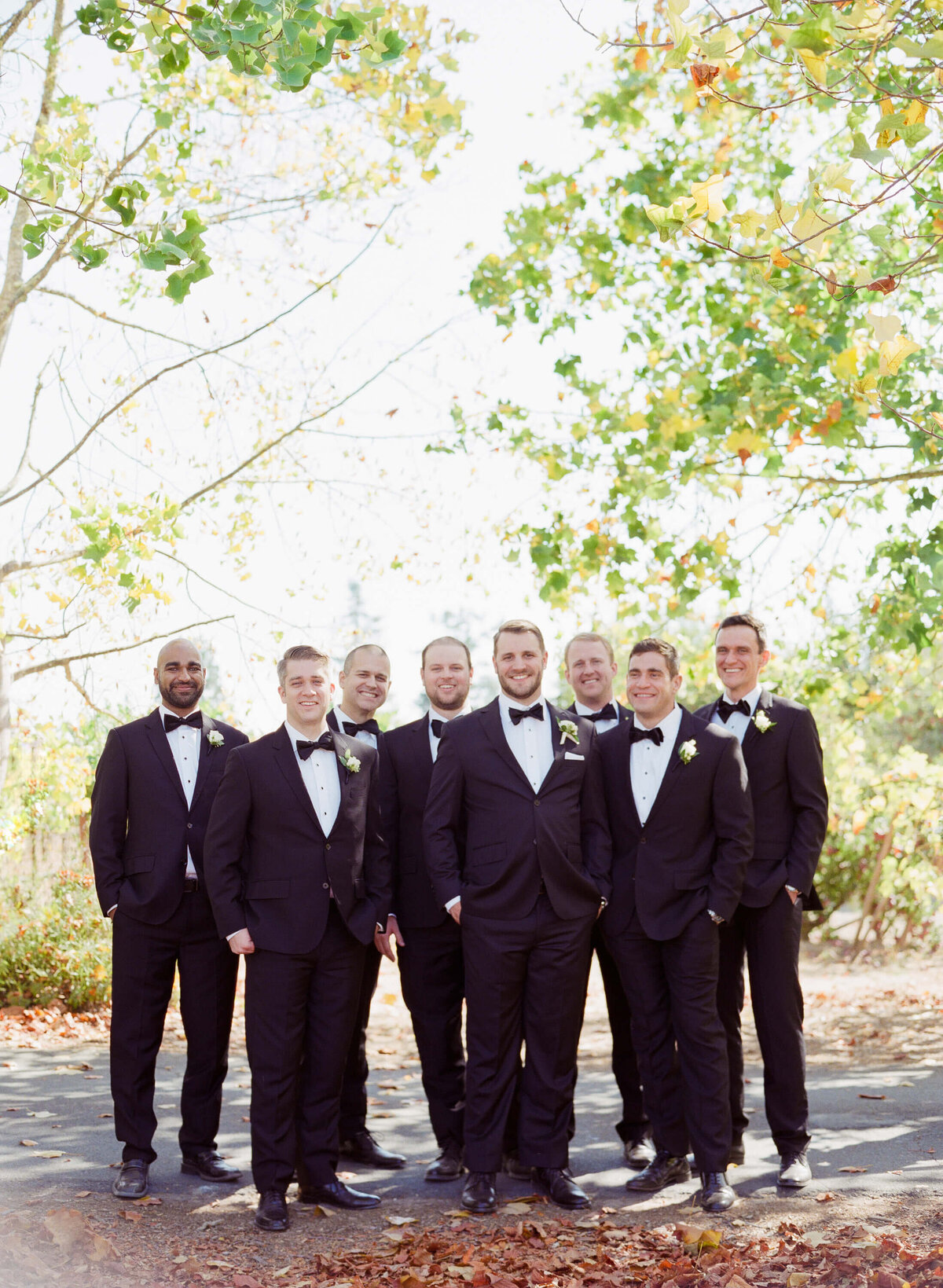 Groom-to-be poses with his best men in suit and bow tie under beautiful green trees and dried autumn leaves.