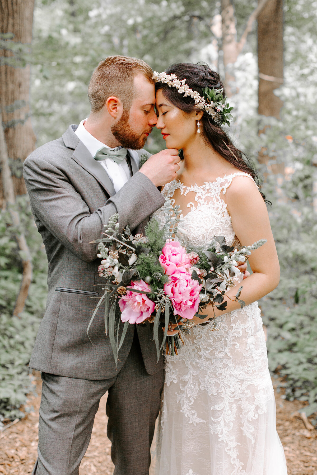 A bride and groom are nose to nose as the both close their eyes, the groom has his hand under the brides chin. They are standing in the woods