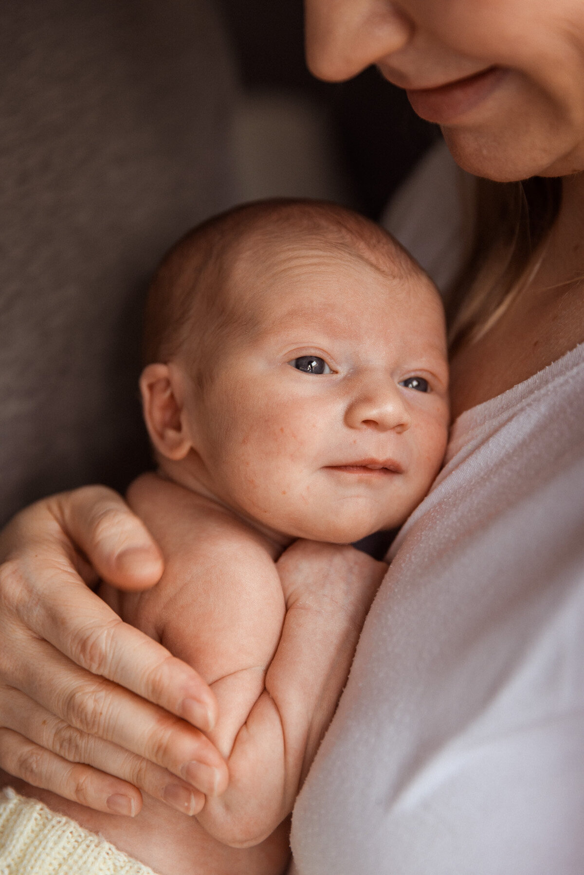 Livsstilsfoto nærbilde av baby som blir holdt mot mors bryst. Man ser hånden til mor på babyens rygg og babyen ser forbi kamera.