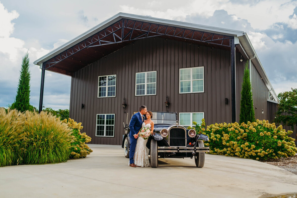 A photo of a bride and groom leaning on a vintage car in front of a Tennessee wedding venue