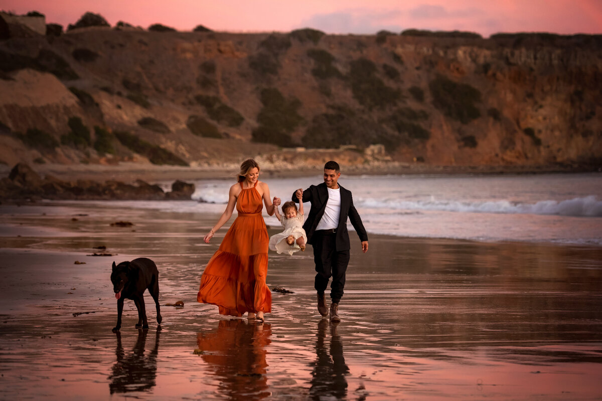 Happy parents Valderrama walking on the beach during pink sunset with daughter and black dog