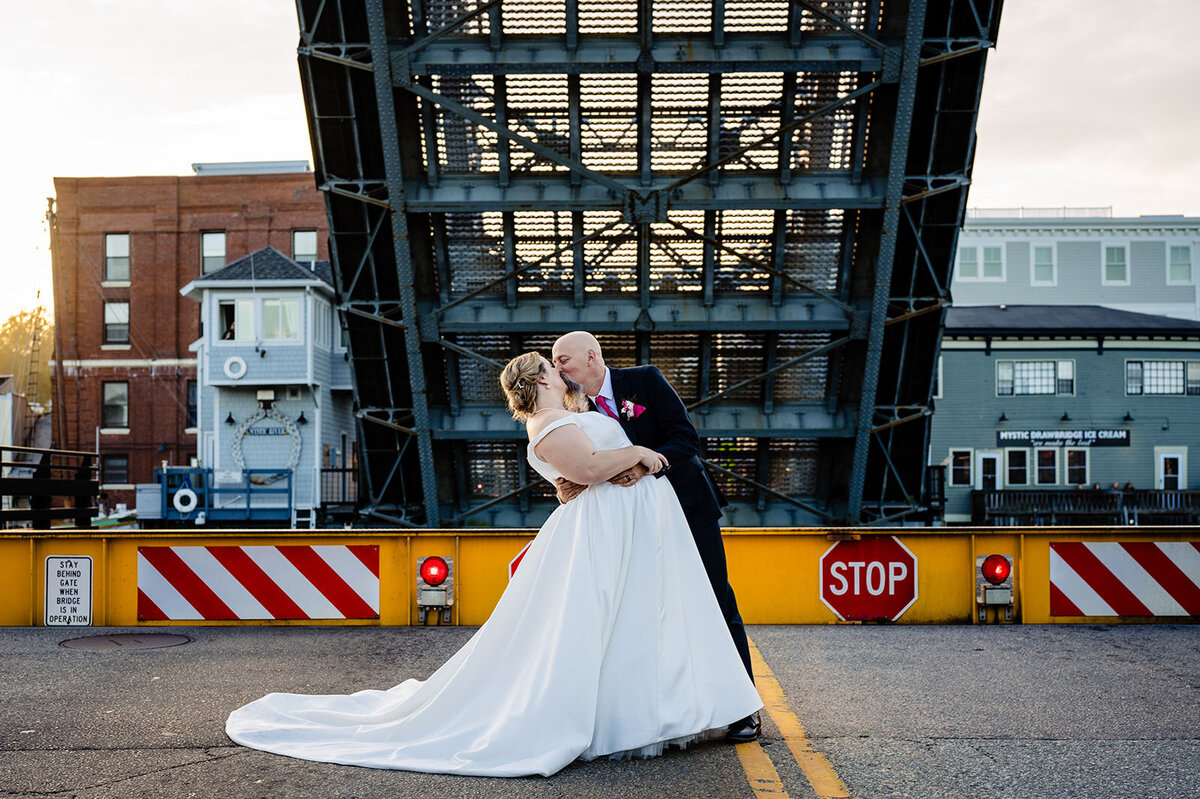 A bride and groom share a romantic moment in the middle of a street with an open drawbridge in the background during sunset