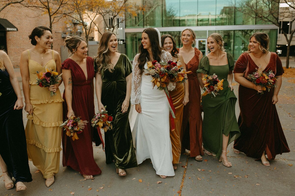 A bride and her bridesmaids in colorful dresses, holding bouquets and smiling as they walk together on a city street during a wedding coordinated in Iowa.