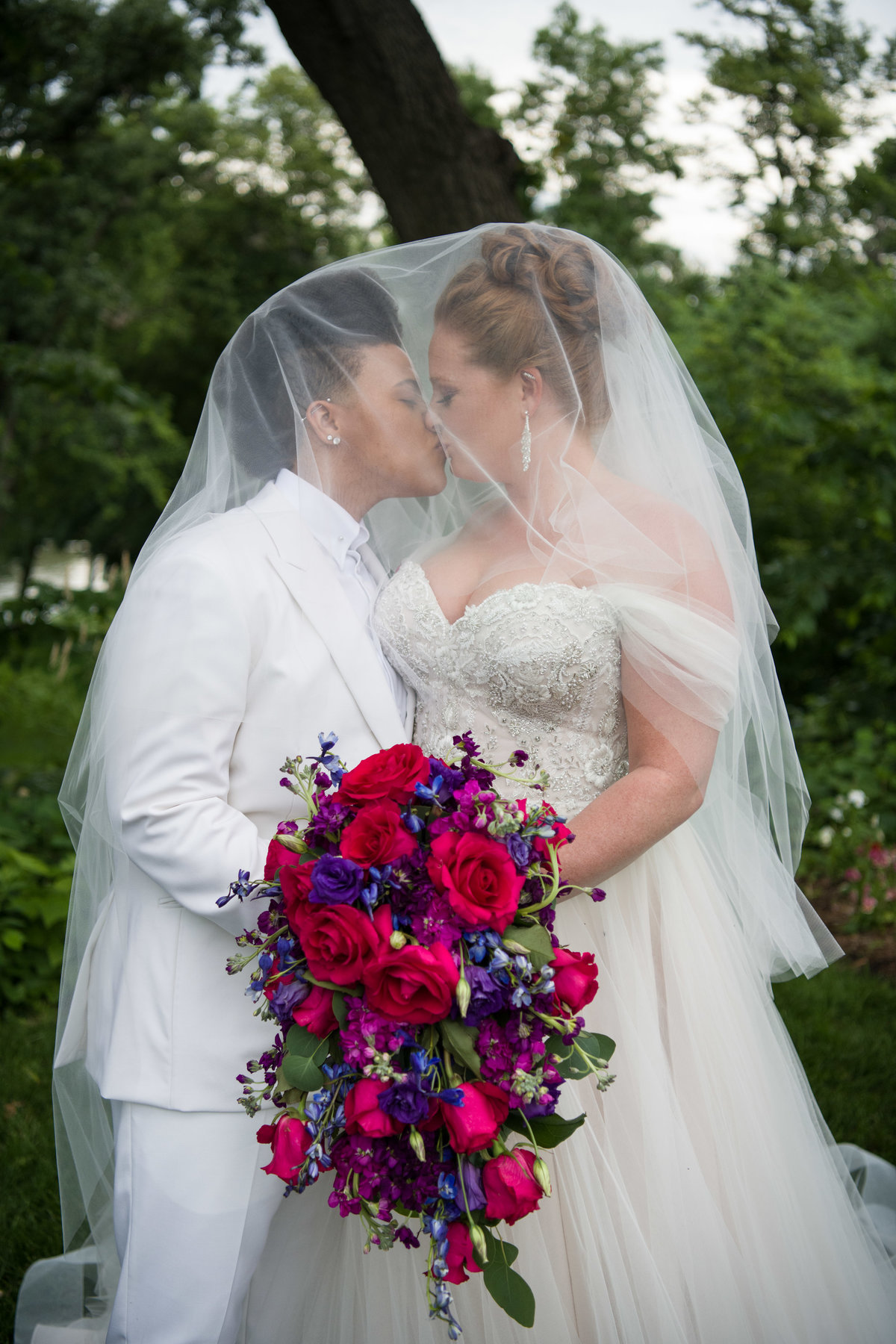 Brides kiss under veil while holding roses.