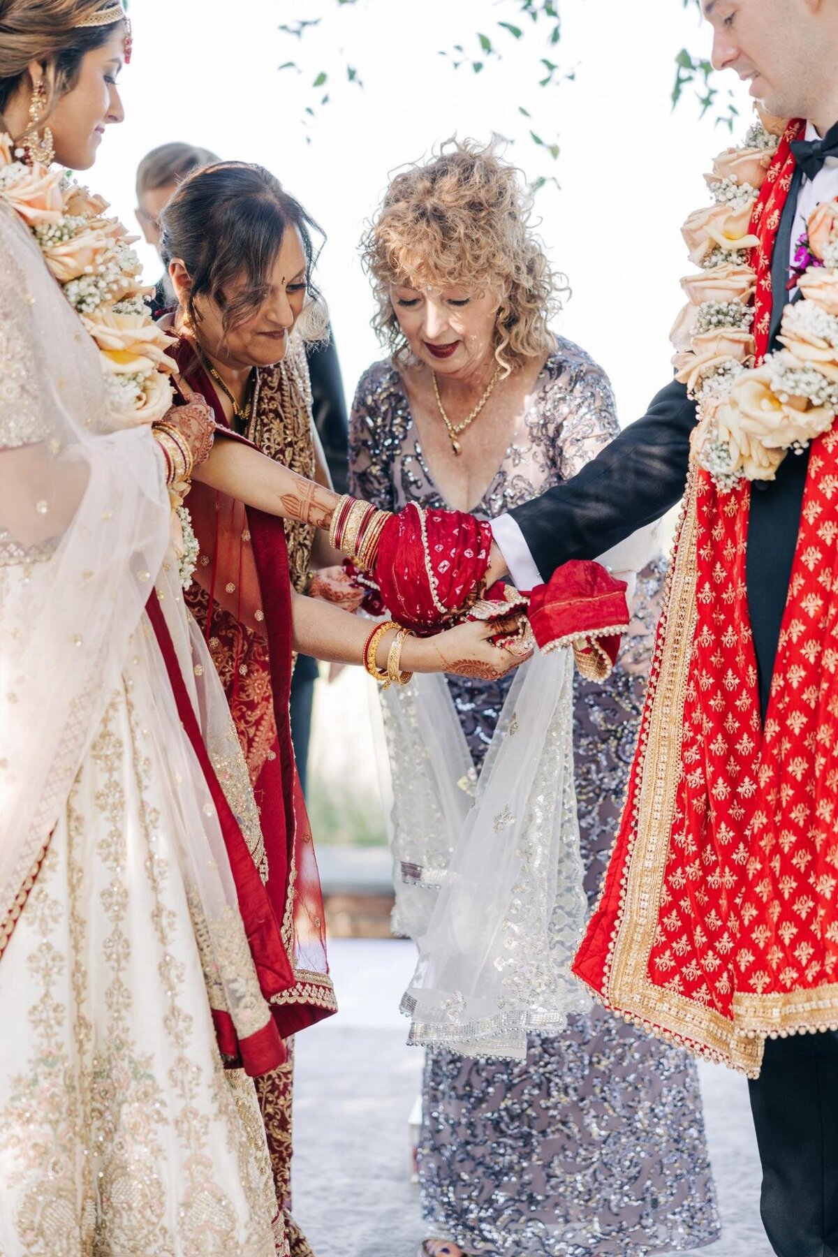 A multicultural wedding ceremony with a bride in a red and white sari receiving blessings from an older woman in a silver dress.