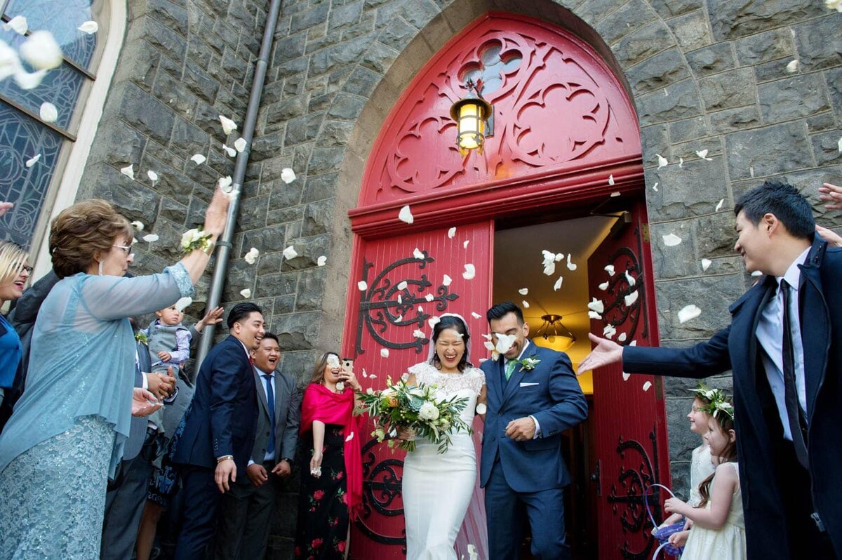 guests toss flowers on a bride and groom as they exit a bright red door of a cathedral