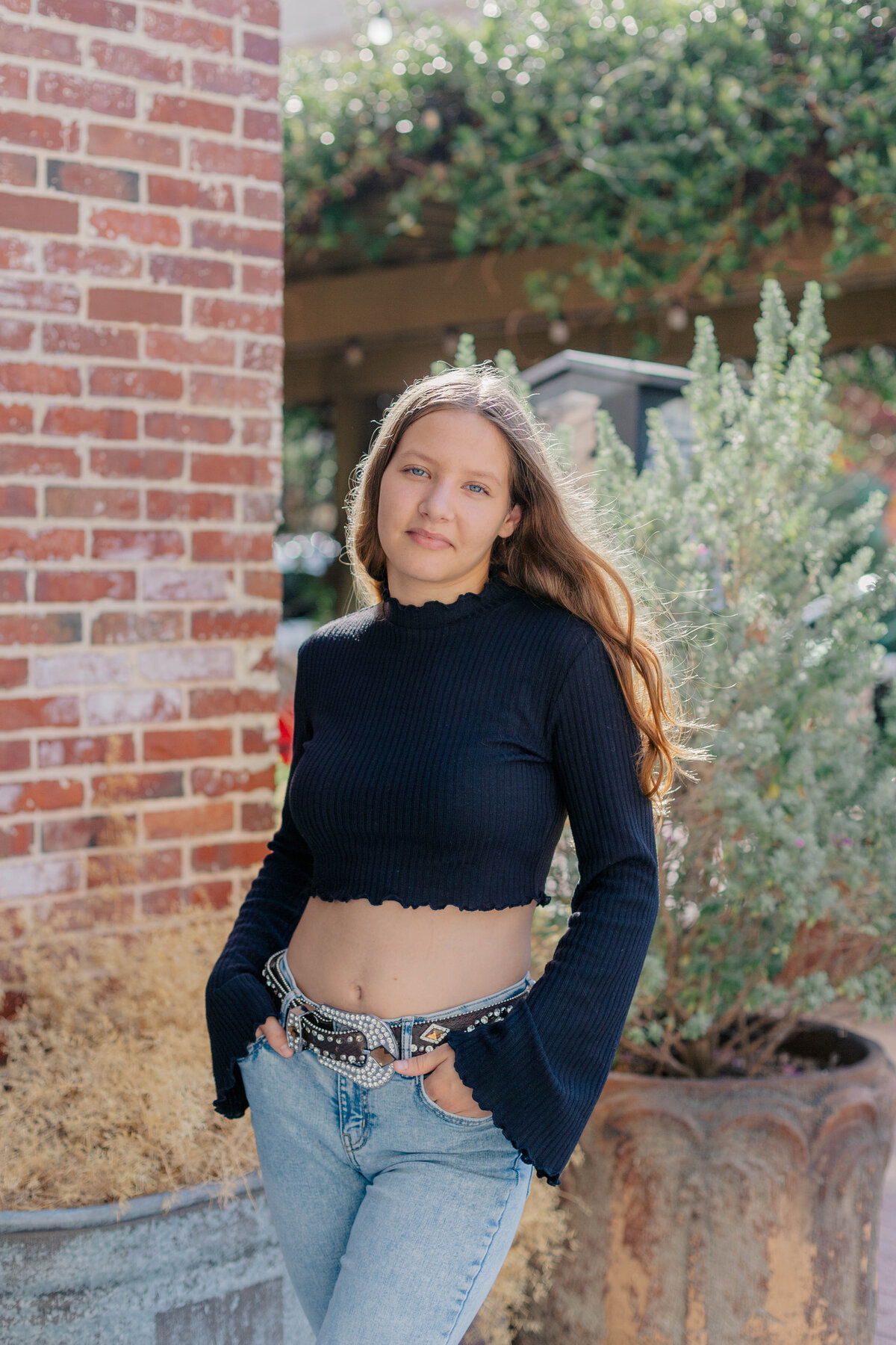 Senior girl stands in a black long sleeve crop top and jeans with a bejeweled belt with her hands in her pockets