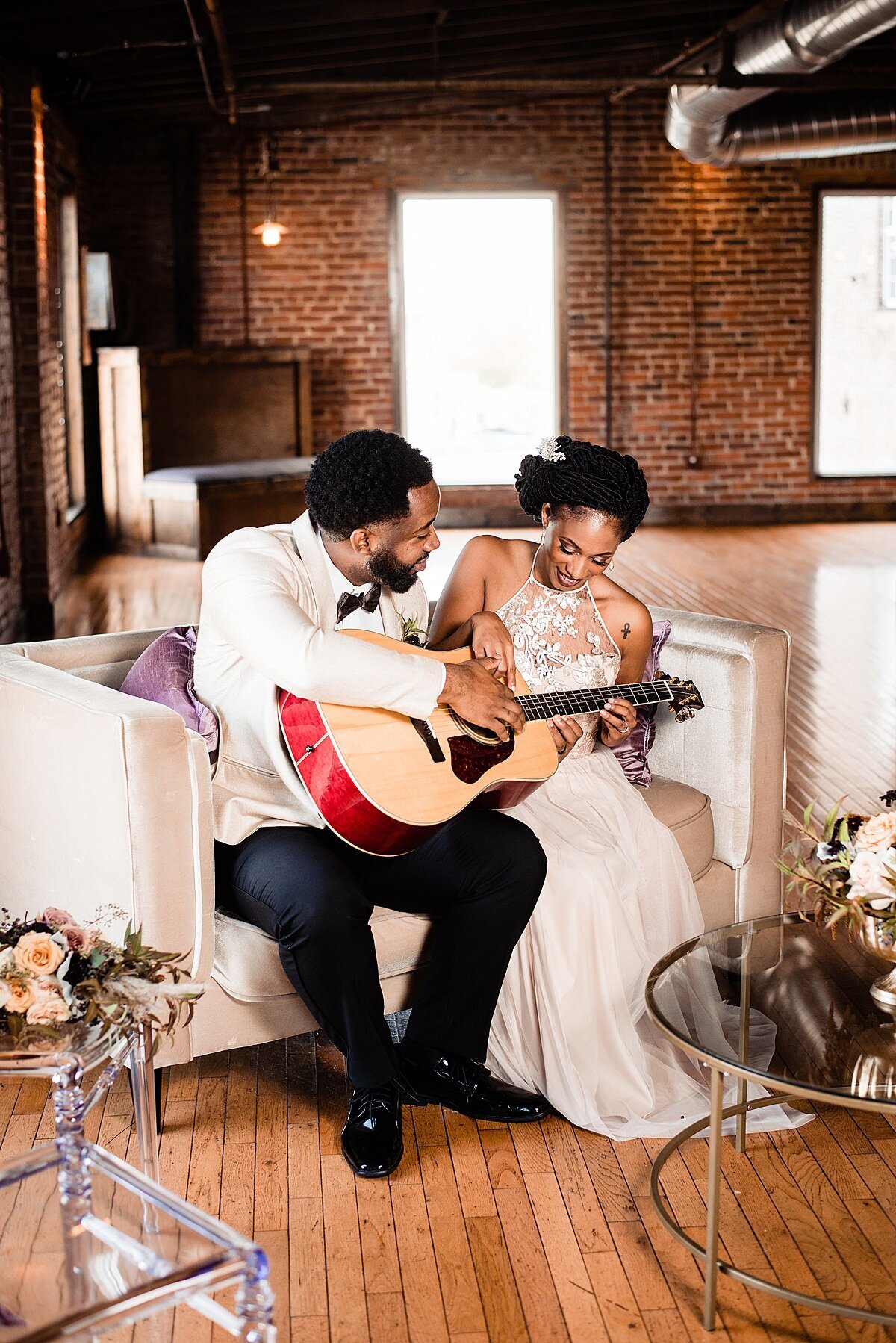 The bride and groom sit in an exposed brick space with hand wood floors and clear glass coffee tables with metal accents and floral bouquets on each table. They are sitting on a white couch as  the groom shows the bride how to play guitar. The groom is wearing a white tuxedo jacket with a white shirt, black bow tie and  black pants. The bride is wearing a halter neck lace wedding dress dress with a  long flowing skirt.