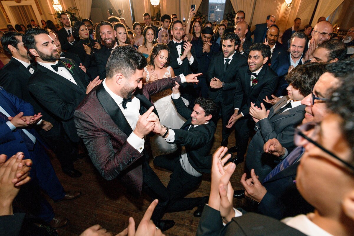 A lively wedding dance floor with guests dressed in formal attire. A groom and a man in a suit enthusiastically dance at the center, surrounded by a circle of guests clapping and cheering. The atmosphere is joyful and celebratory.