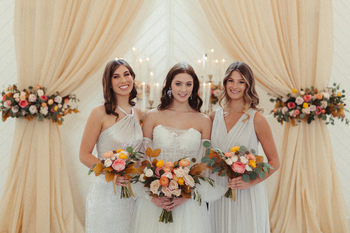 A bride wearing a white wedding gown poses with bridesmaids wearing light gray gowns with bouquets in hand.