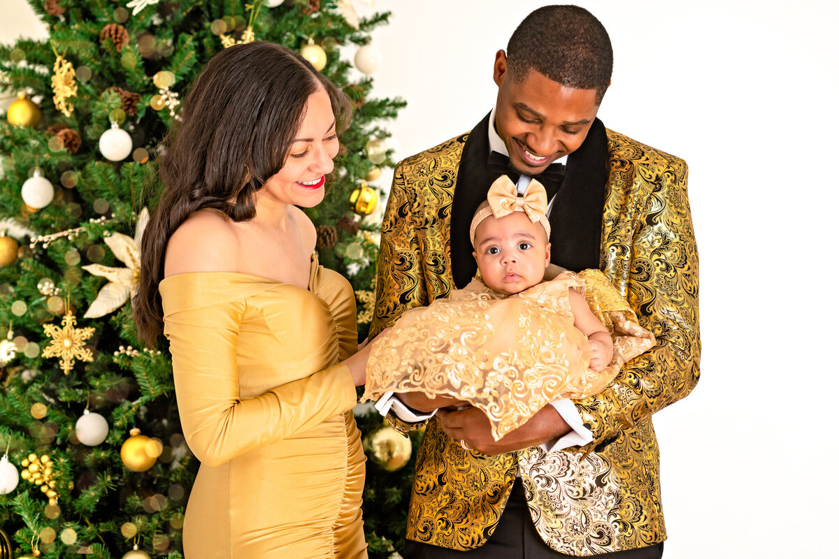 A mom and dad in formal attire are looking down at their baby girl as they pose for Christmas pictures  with Ashley Zohil Photography.