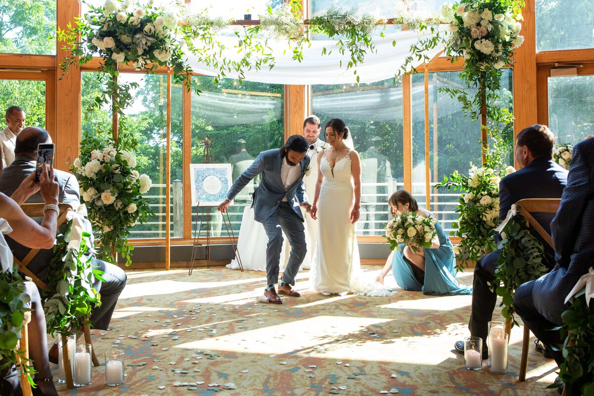 A wedding couple watches a guest perform a dance move in a sunlit venue with large windows and floral decorations during an event in Davenport.