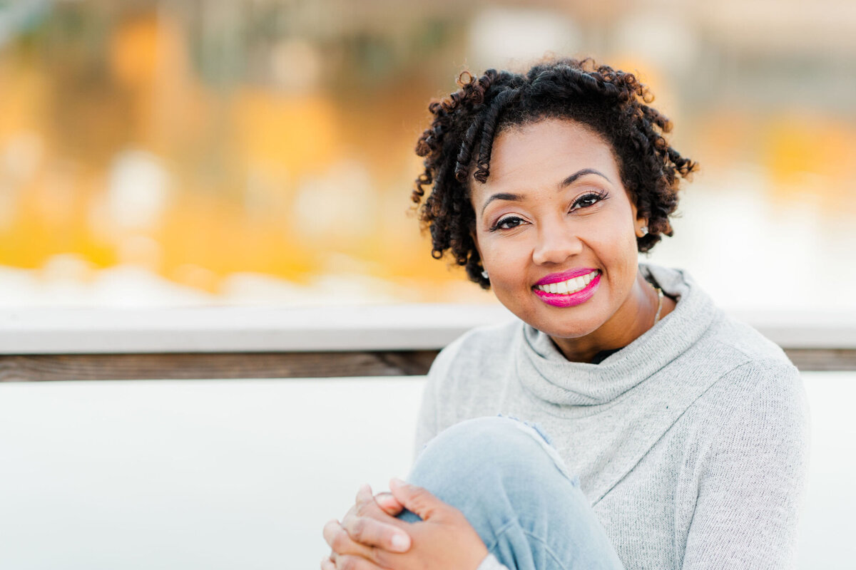 Woman in a grey sweatshirt smiling at the camera with fall colors in reflecting off  the lake