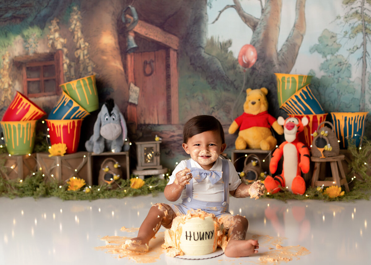 Modern Winnie the Pooh themed cake smash. Baby boy in a white shirt, blue overalls and a bow tie sitting in front of a hunny pot cake. The baby is covered in icing all over his legs and face. In the background, there is Pooh's house, buckets of honey, and pooh, tigger, and eeyore stuffed animals.