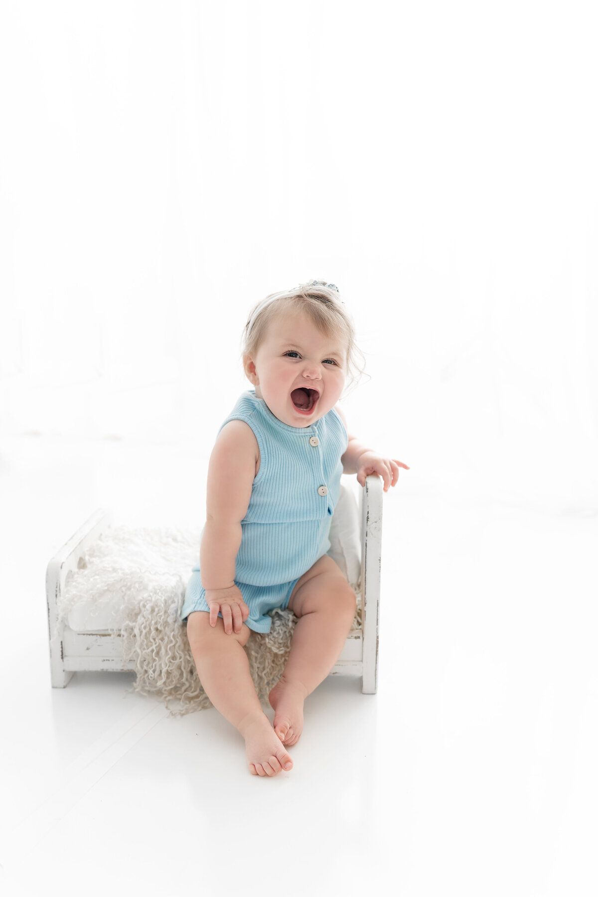 A toddler girl yawns big while sitting on a tiny wooden bed in a blue onesie
