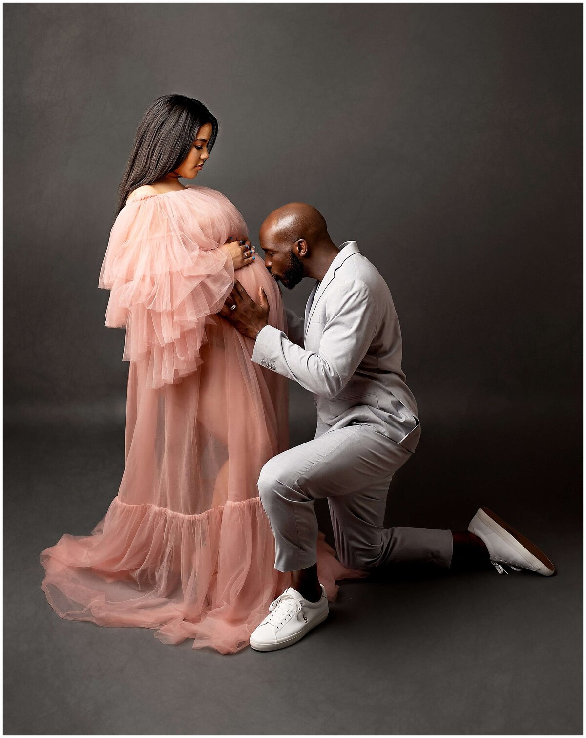 A couple in a maternity photoshoot, with the father kneeling down and tenderly kissing the pregnant belly of his partner. The intimate moment captures the love and anticipation of the couple as they await the arrival of their baby.
