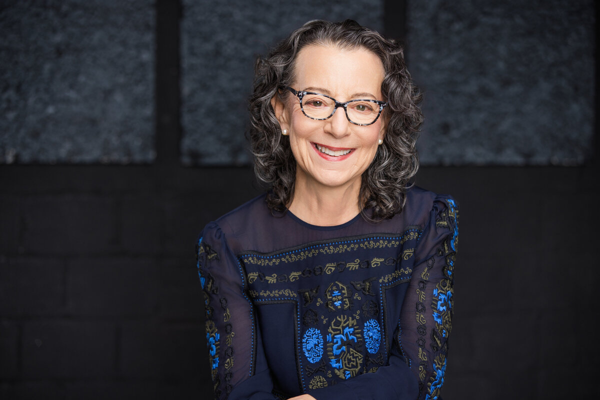 Professional headshot of an older Caucasian woman with curly gray hair and glasses, captured in Cincinnati for personal branding. She wears a stylish navy blue top with intricate patterns, enhancing her friendly and approachable demeanor. Perfect for updating professional profiles, LinkedIn, and personal branding materials. Optimize your Cincinnati headshots with expert photography services for a polished, professional image.