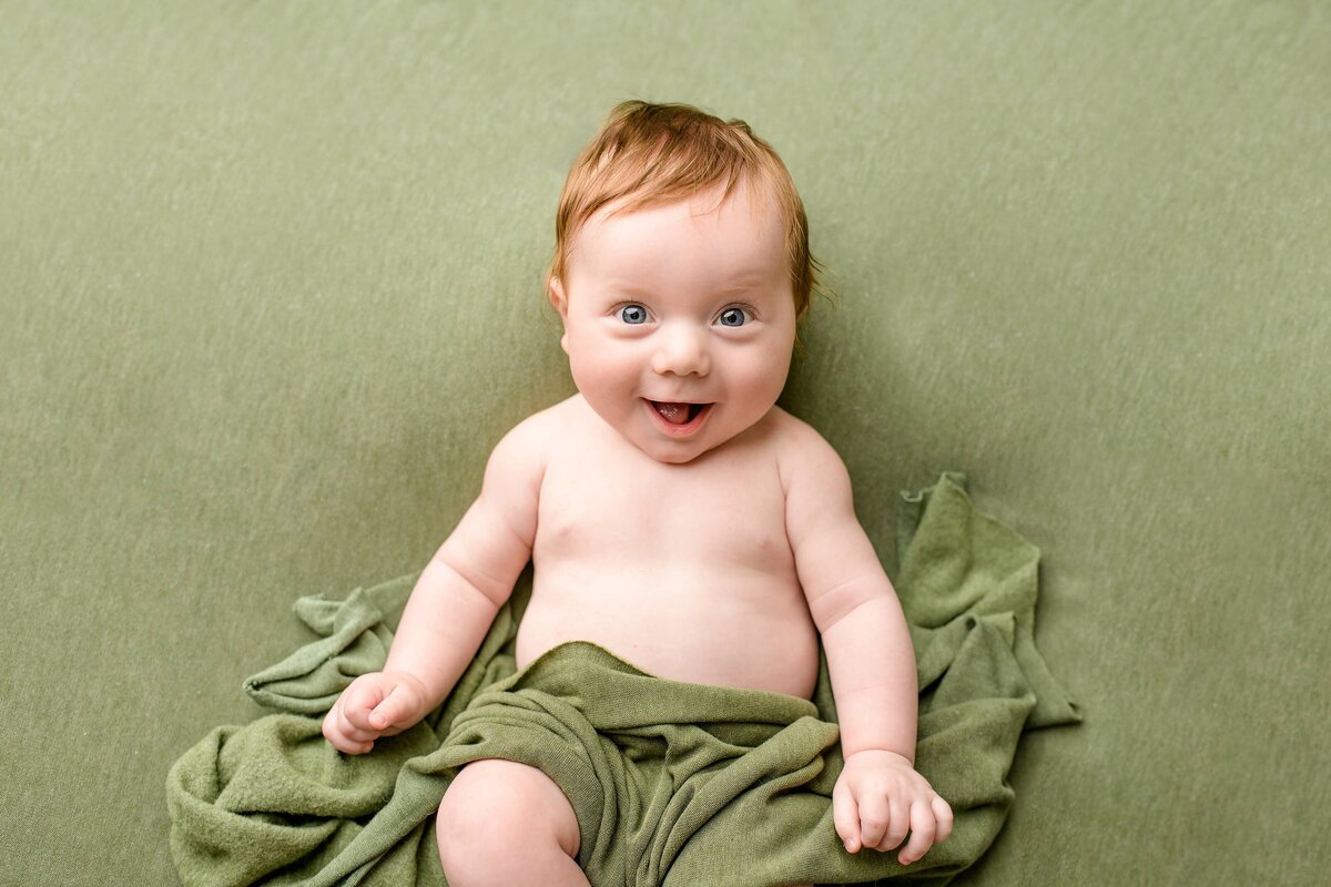 3 month old baby boy from the Charlotte area with red hair and big blue eyes Smiles as he lays on a green background during his Milestone session