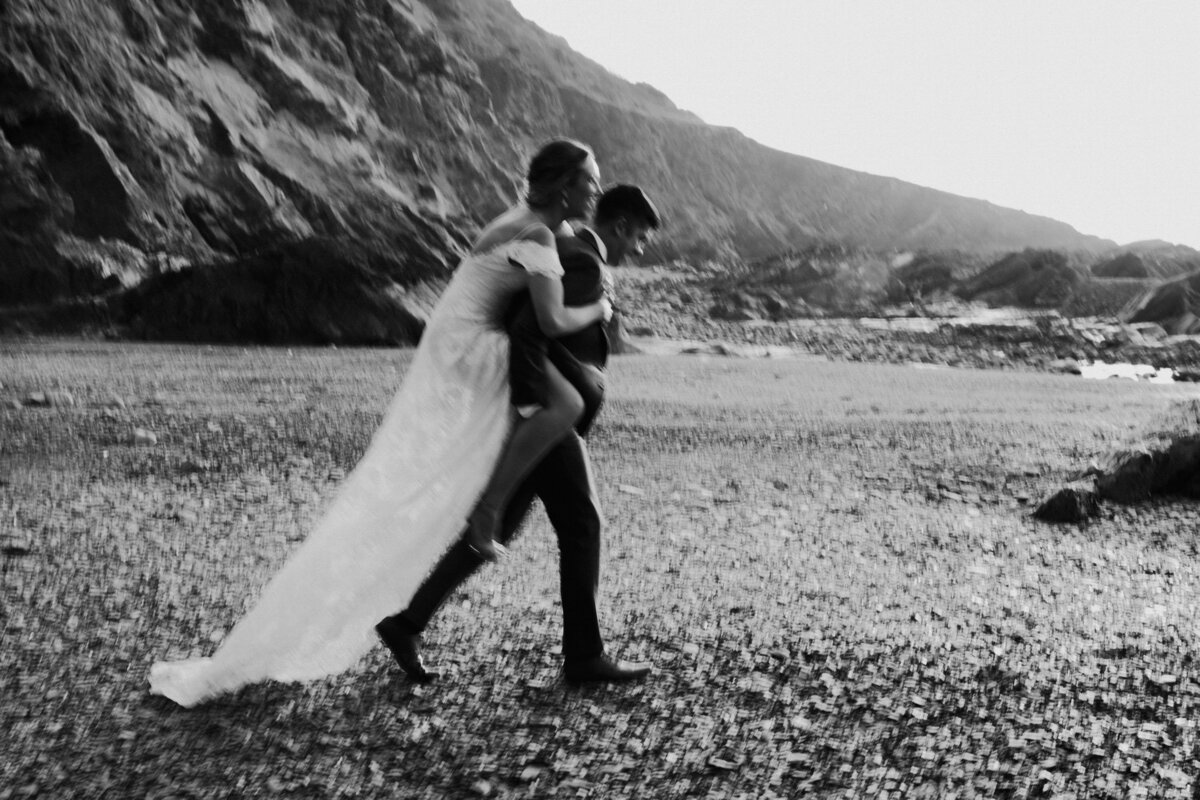 Black and white photo of groom carrying bride on coast