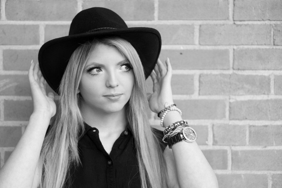 Girl with long blonde hair and a black cowboy hat stands in front of a brick wall