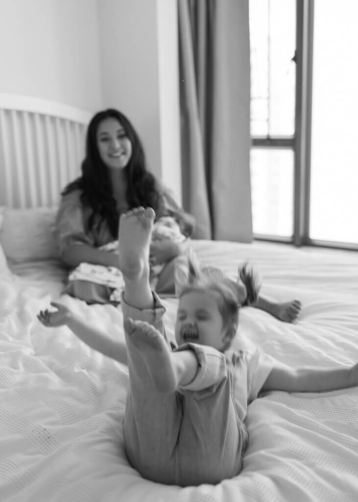 Black and white photo of toddler playing on the bed while mother smiles holding her newborn in the background.