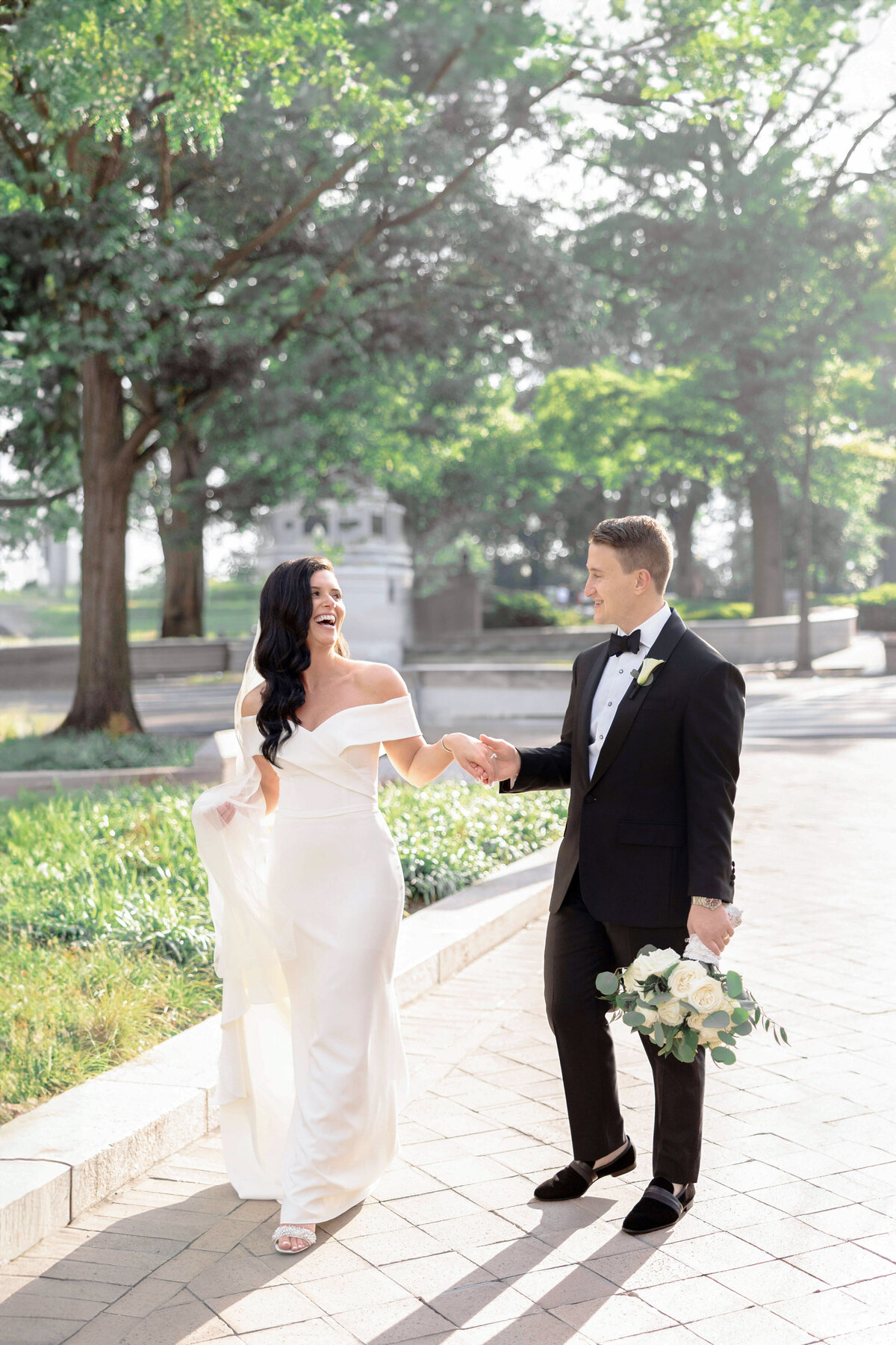A couple dressed in wedding attire walks hand in hand outdoors. The bride wears a white gown and the groom wears a black tuxedo. They both smile joyfully while surrounded by greenery and sunlight.