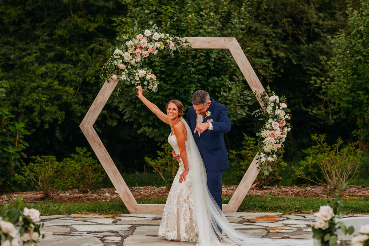 Photo of a bride dancing with the groom hugged her and pointed at her while standing in front of an arch