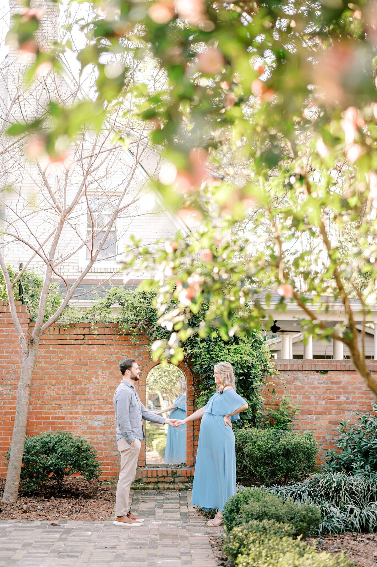 An expecting couple stand facing each other while holding hands in front of the  garden wall at Wing Haven Estate.
