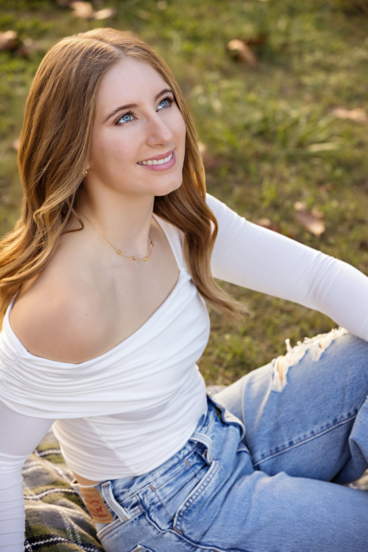 Close-up portrait of a high school senior in jeans and a white top looking up and away from camera sitting on a blanket in an outdoor setting