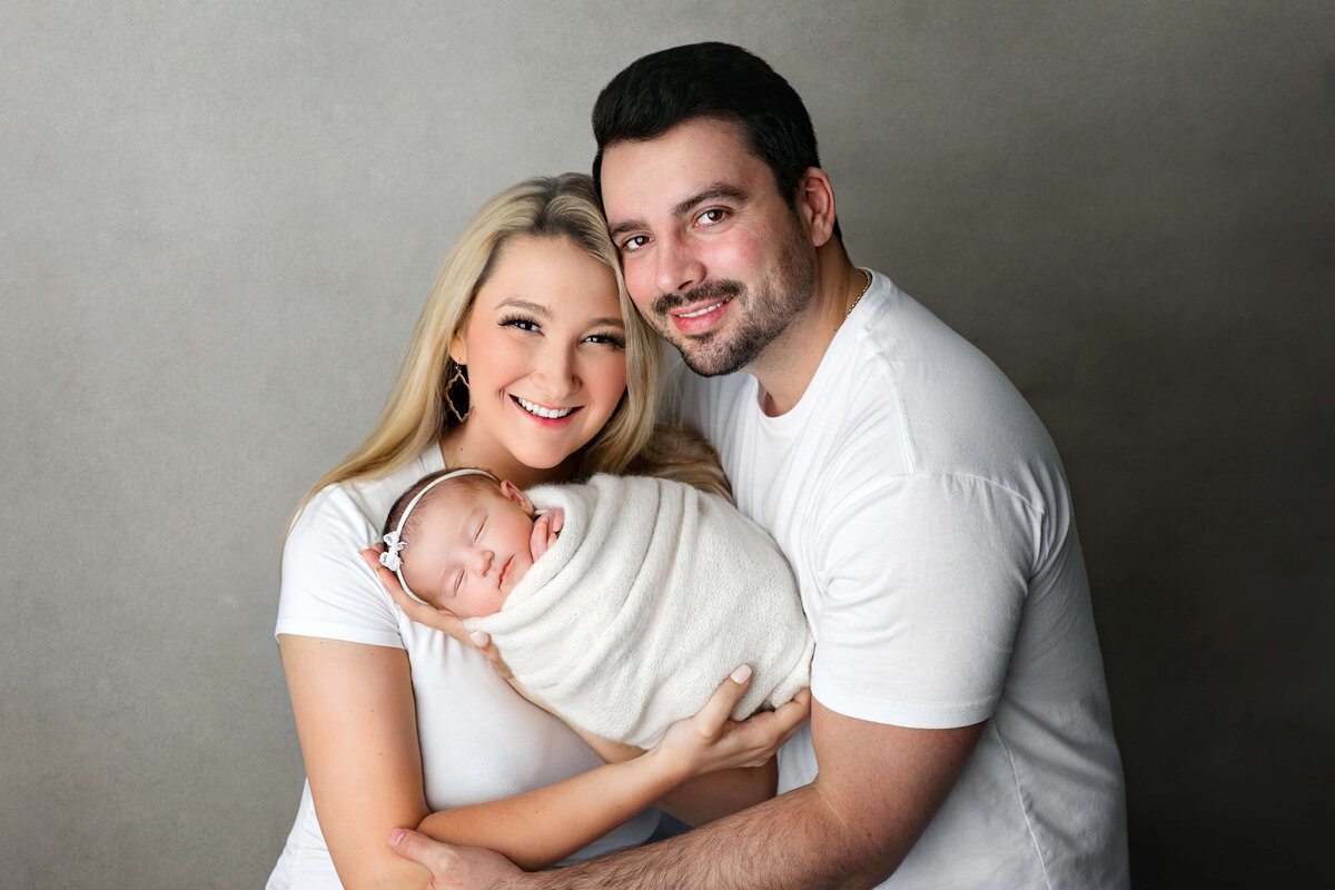new mom and dad holding their newborn baby girl wearing  white on a grey backdrop at their VA Newborn  Photoshoot