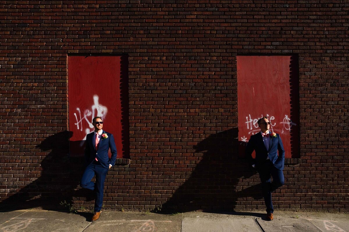Two grooms leaning against a brick wall.