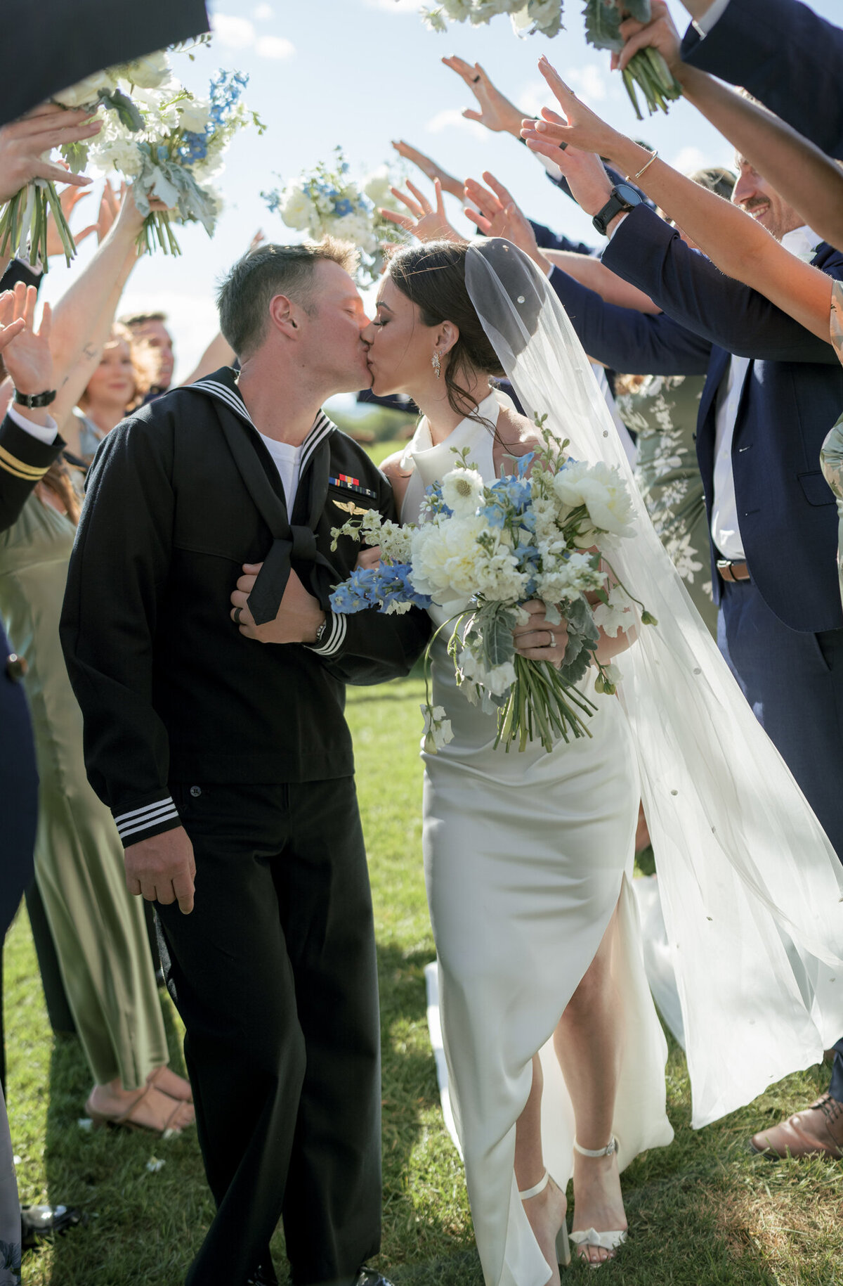 Bride and groom share a kiss beneath a joyful arch formed by their bridal party’s raised arms, set against Teton Mountains of a French blue summer wedding in Jackson, WY.