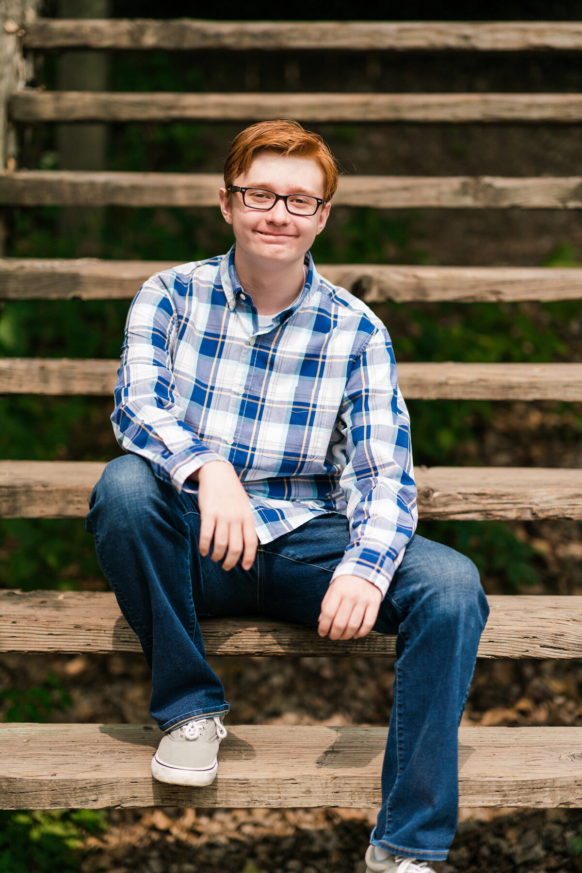 Senior boy dressed in flannel and jeans sits on stairs.