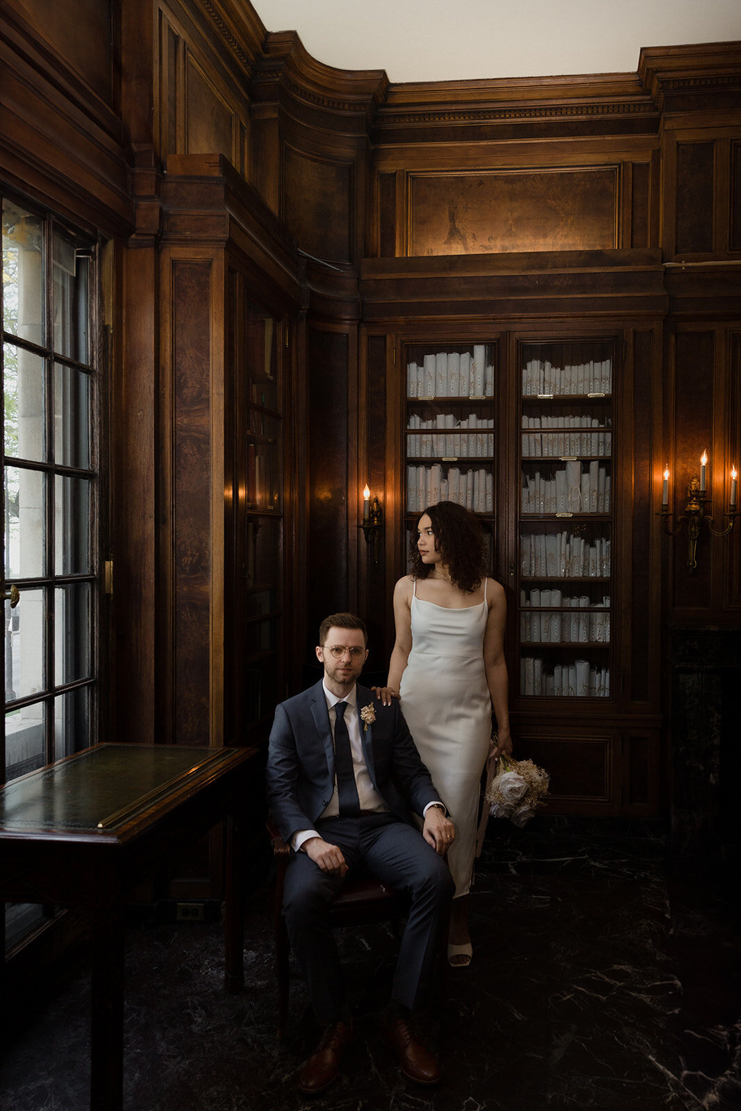 Just Married photo session couple in dark wood library with large window. Grooms sits in leather chair and looks on while bride stands next to him with her hand on his shoulder and looks out the window.