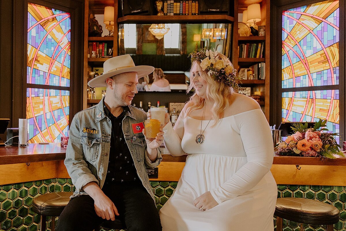 The boho bride, wearing a long sleeved dress and flower crown clinks glasses with the groom as they sample their signature cocktails. The groom, wearing a black shirt and black  pants with a Denim & Spirits custom denim jacket and cowboy hat. The bride and groom sit at the green tiled bar in front of the stained glass windows at Rosemary & Beauty Queen in Nashville.