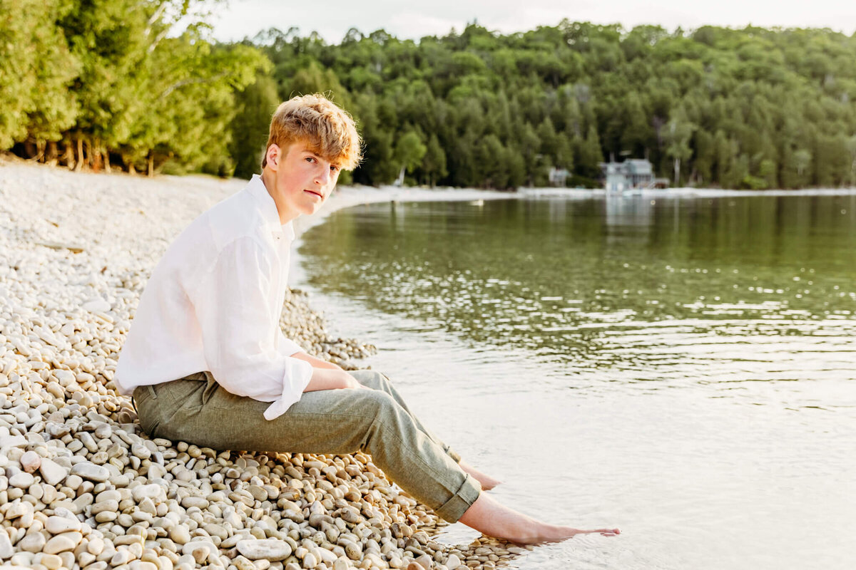 handsome teen boy sitting on a rocky beach with his feet in the water for his senior photography experience