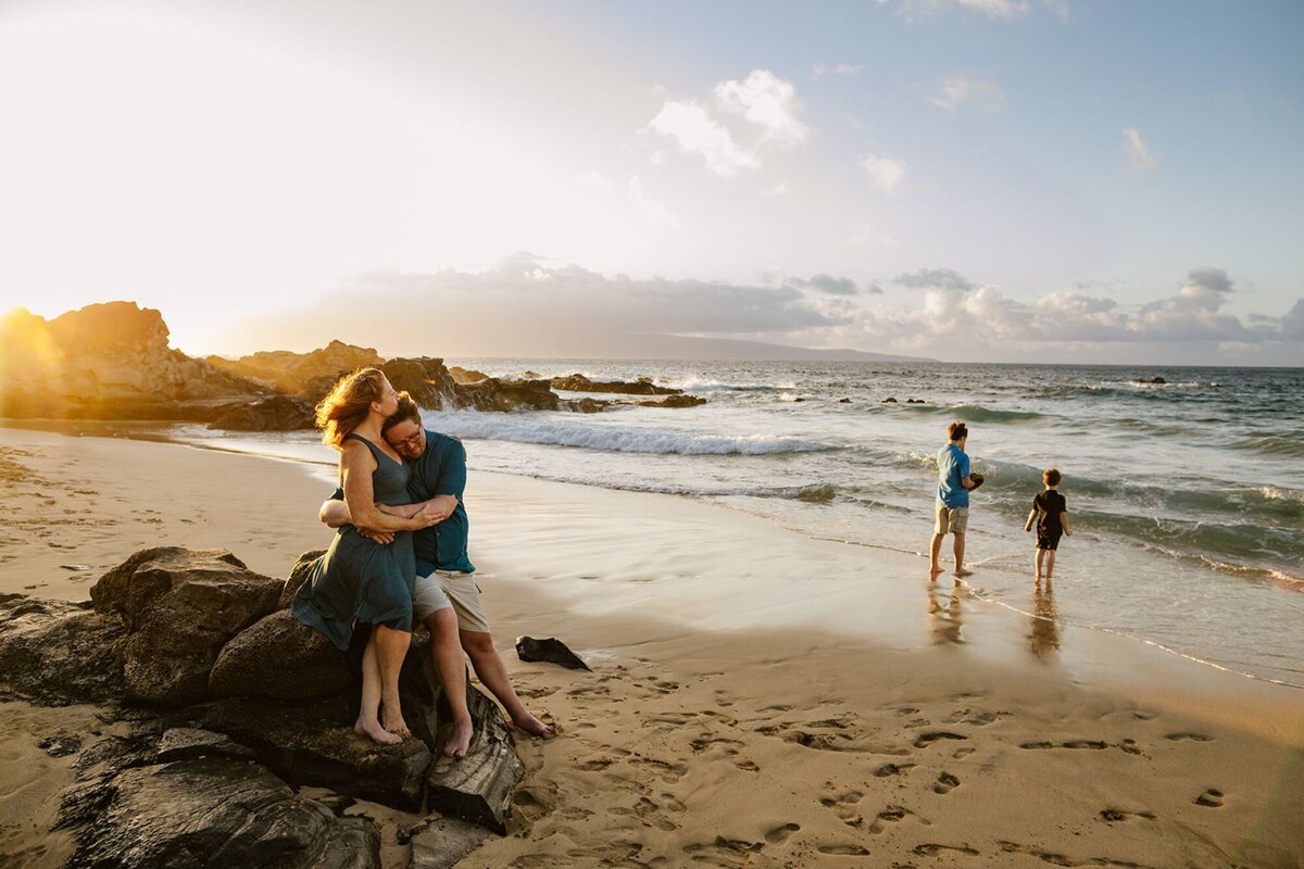 A man and woman hug on a rock as their two boys play in the ocean in the background.