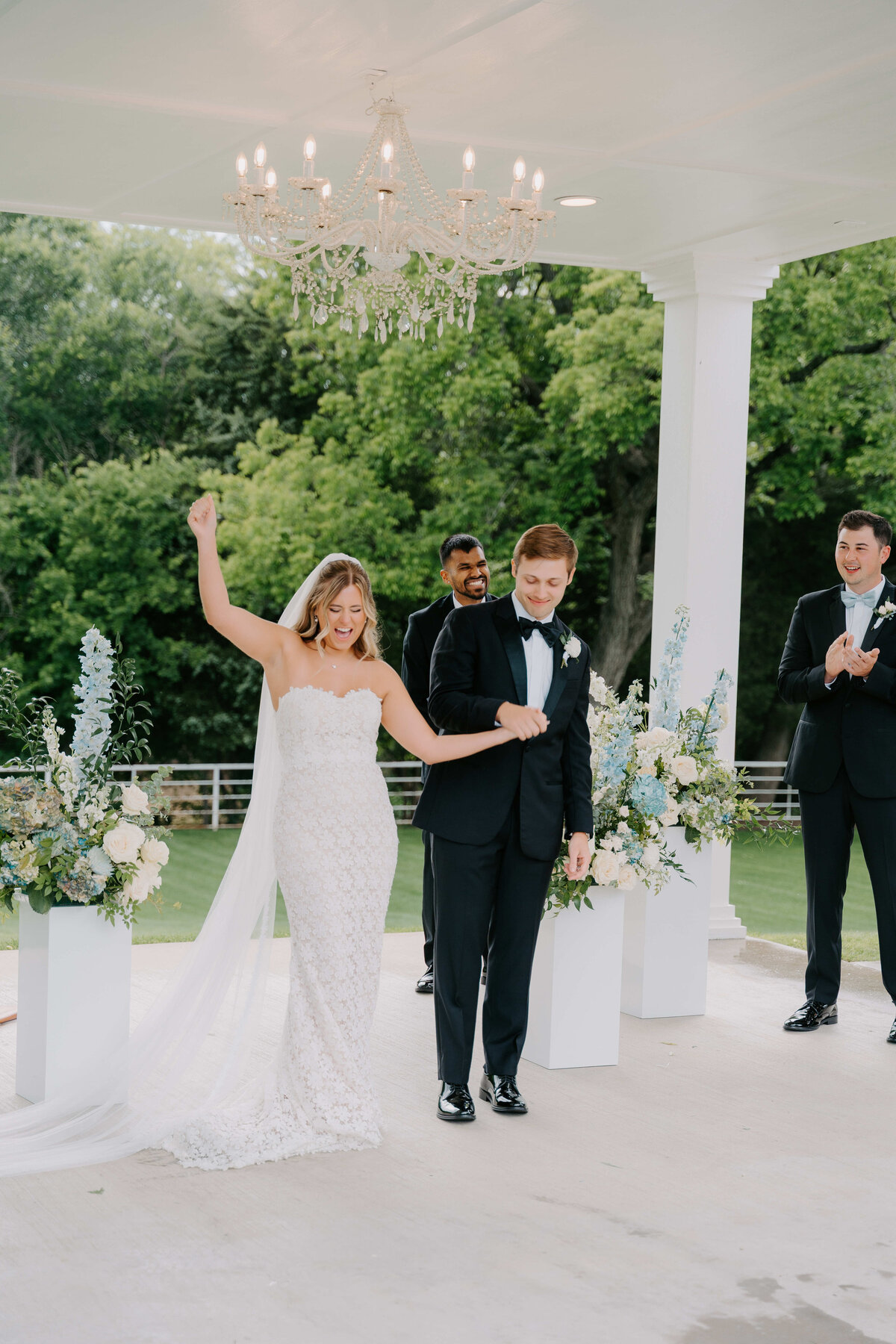 A joyful bride and groom, in a white dress and black tuxedo, celebrate under a chandelier on a veranda, surrounded by bouquets and clapping guests.