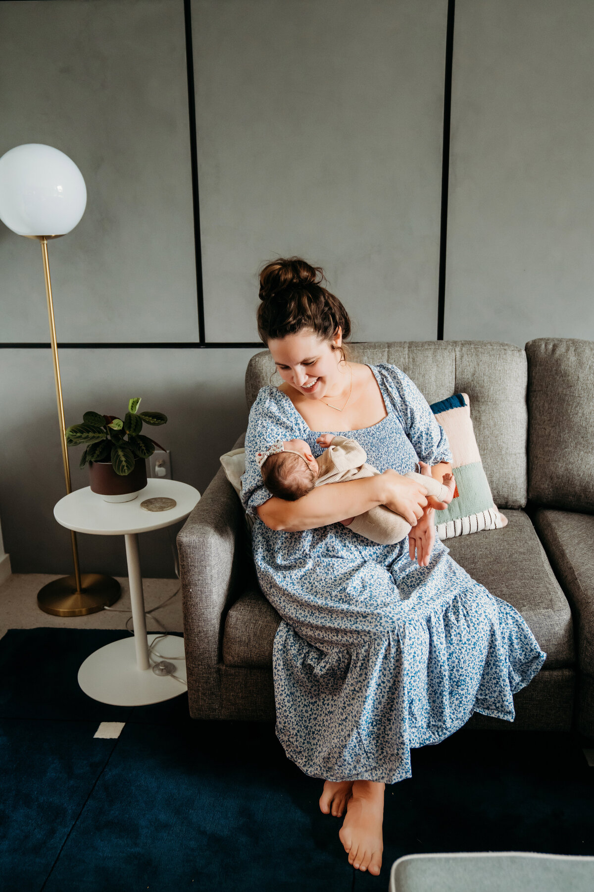 Newborn Photographer, a mother sits on couch at home and holds her baby in her arms smiling
