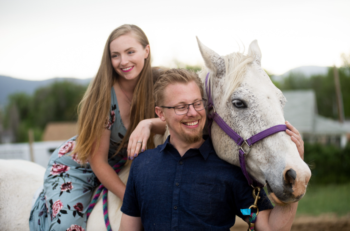An engaged couple poses with their white horse. The woman sits on the horse leaning forward and smiling. The man stands to the side of the hose, petting its head.